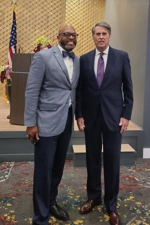 Two men wearing suits pose for a picture while smiling.